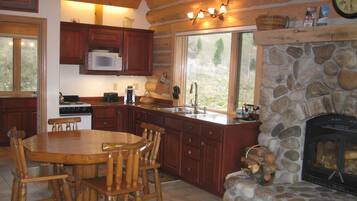 A log cabin-style kitchen with a stone fireplace, wooden cabinetry, and a dining area with a wooden table and chairs.