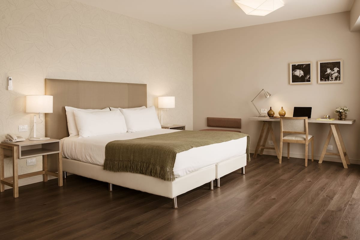 A modern hotel room with a large bed, bedside tables, a desk with a chair, and a lamp.