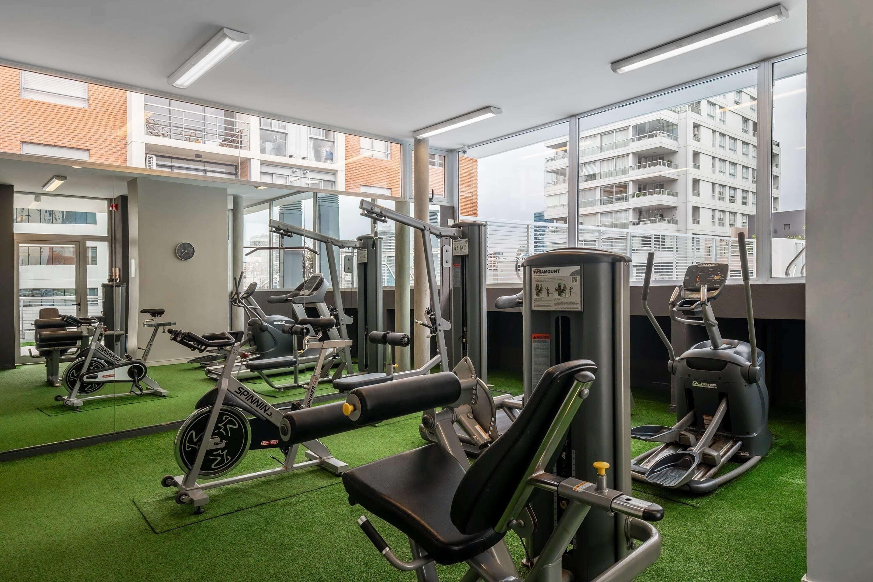 Uma academia com diversos equipamentos de exercício, um relógio na parede e vista para um edifício através de grandes janelas.