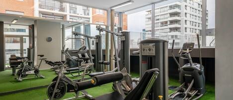 Un gimnasio con diversas máquinas de ejercicio, un reloj en la pared y vistas a un edificio a través de amplias ventanas.