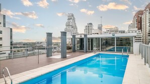 Ein Pool auf dem Dach mit Blick auf die Stadt, umgeben von modernen Gebäuden und blauem Himmel.