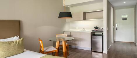 Habitación de hotel con cama, una mesita de vidrio, una silla, una kitchenette y una puerta que da a otra habitación.