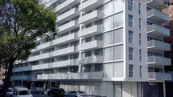 Un edificio residencial de varios pisos con balcones, una zona de estacionamiento y un árbol en la fachada.