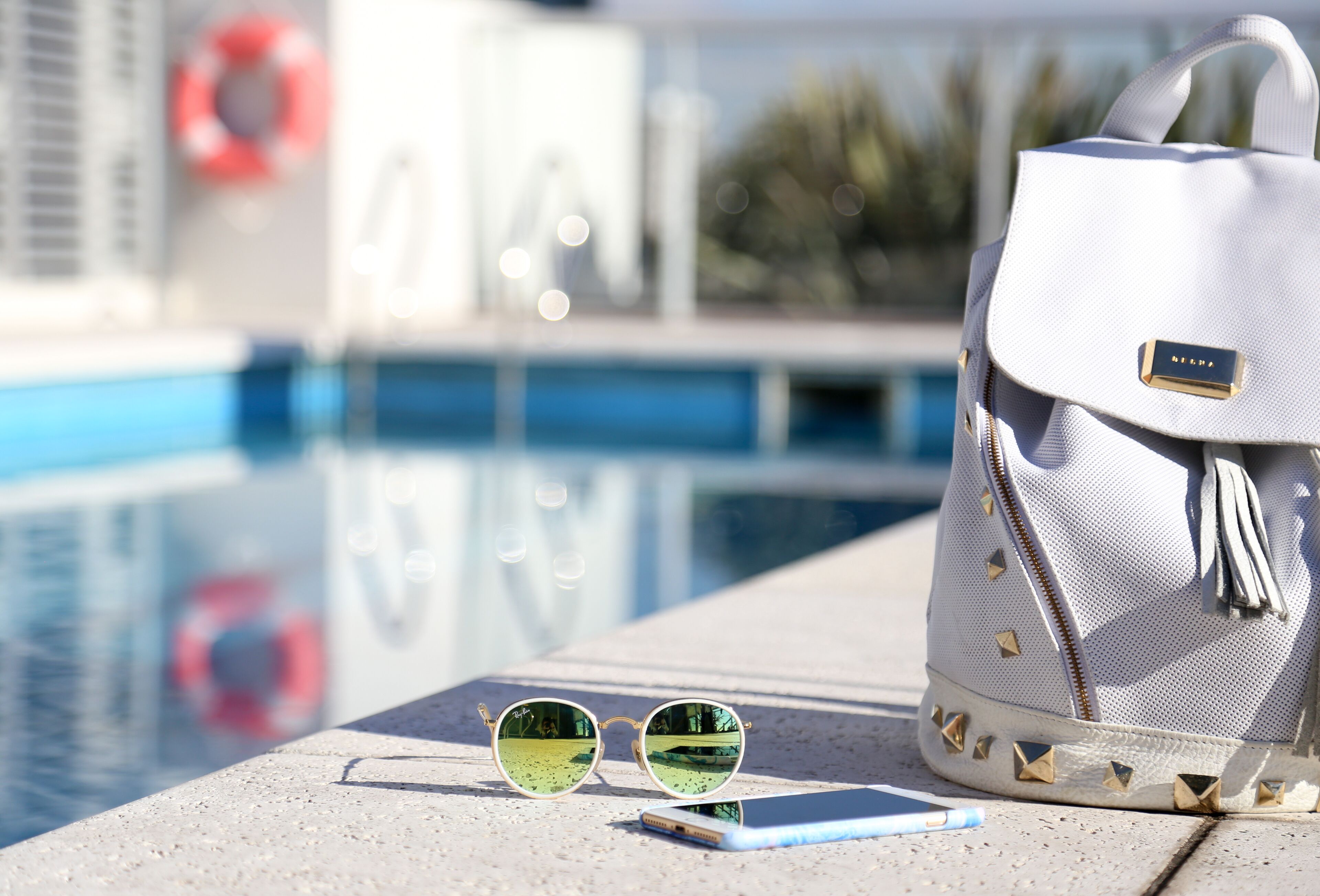 Sunglasses, a smartphone, and a white handbag with a gold zipper detail by a poolside.