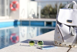 Sunglasses, a smartphone, and a white handbag with a gold zipper detail by a poolside.