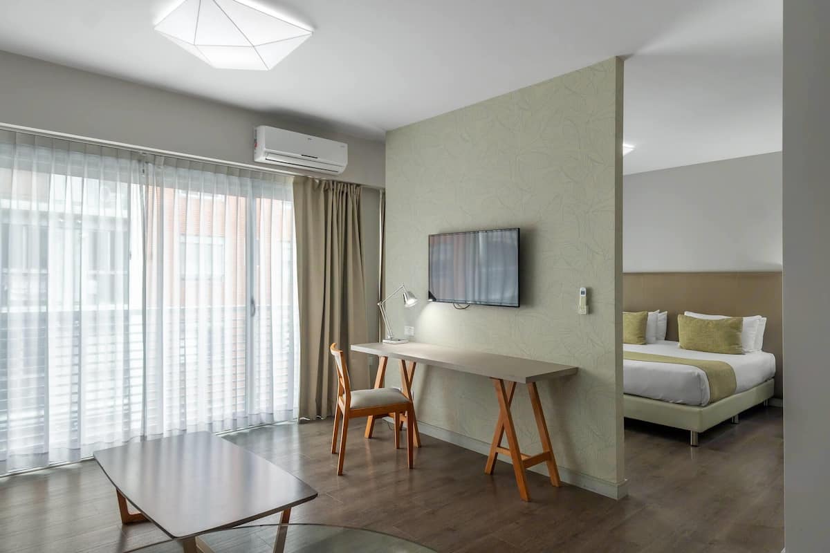 A modern hotel room with a large window, a flat-screen TV mounted on the wall, a bed with white bedding, and a desk with a chair.