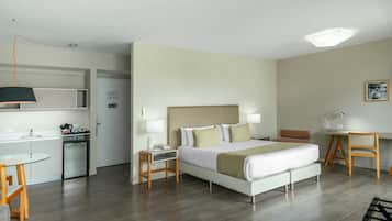 A modern hotel room with a bed, bedside tables, a desk, a chair, a small kitchenette, and a glass table.