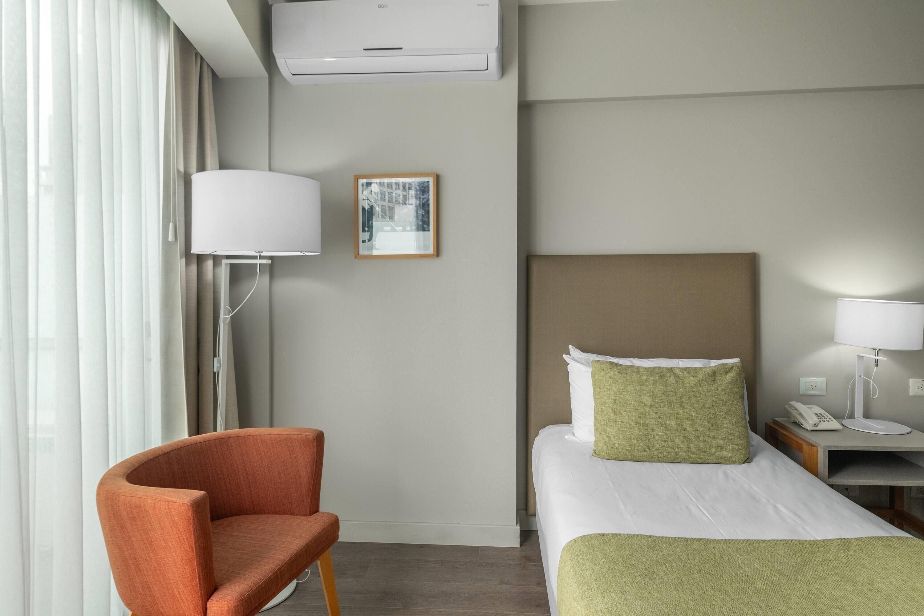 Quarto de hotel com cama, uma cadeira, mesa de cabeceira com abajur, telefone e um quadro na parede.