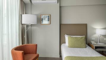 Ett hotellrum med en säng, en stol, ett sängbord med en lampa, en telefon och en tavla på väggen.