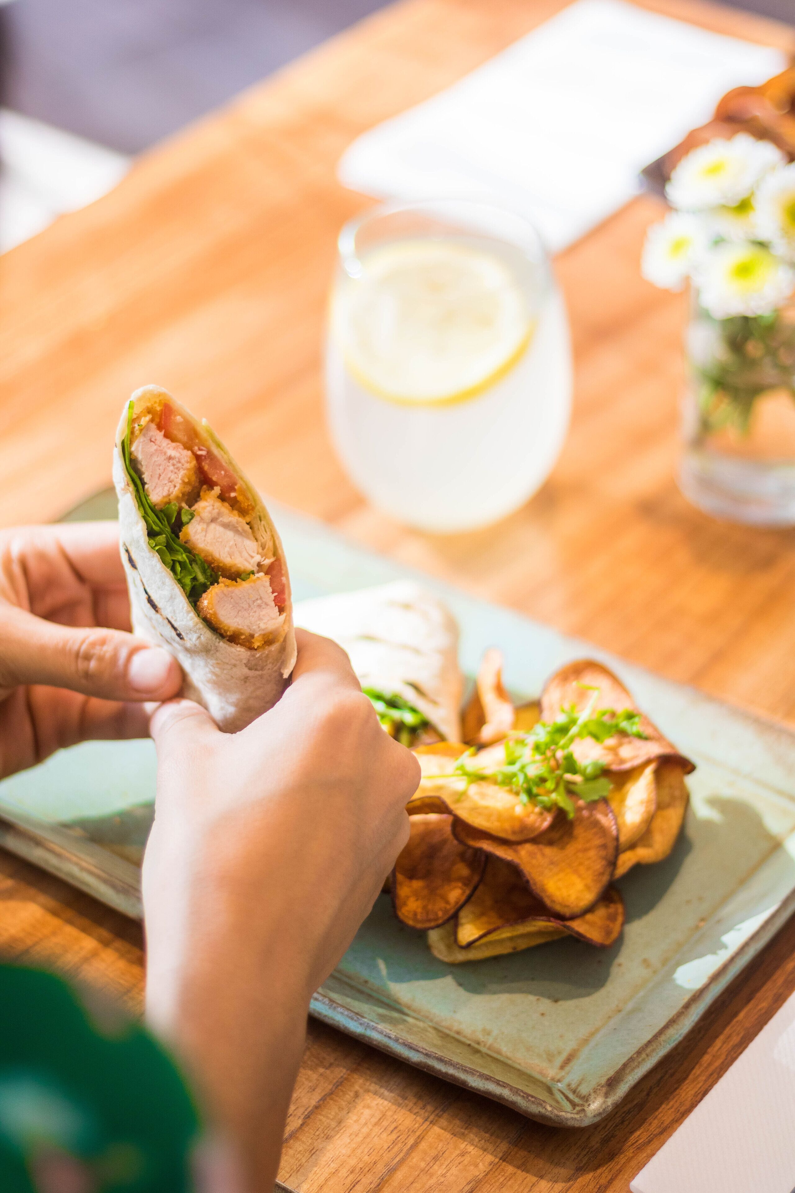 A person holding a wrap with meat and vegetables, a plate of chips, and a pitcher of lemonade on a wooden table.