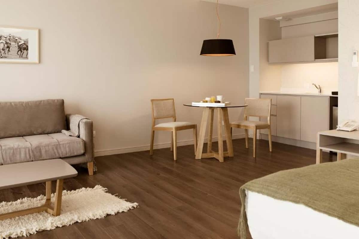 A compact hotel room with a bed, a sofa, a small table with chairs, a kitchen area, and a hanging light fixture.