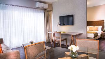 Een moderne hotelkamer met een glazen eettafel, twee stoelen, een flatscreen televisie aan de muur, een bed met hoofdeinde en een raam met transparante gordijnen.
