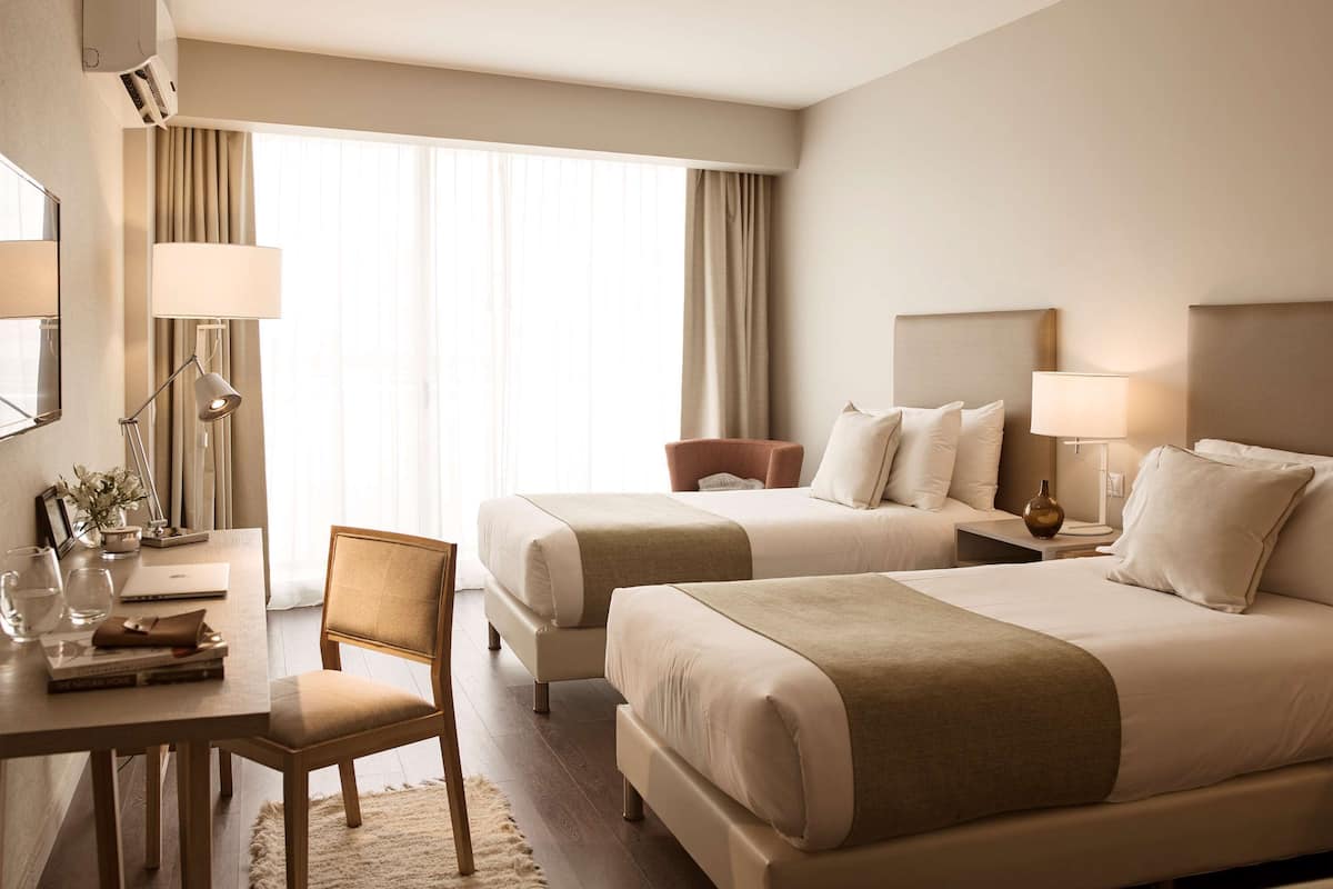 A hotel room with two beds, a desk with a chair, a lamp, and a window with curtains.