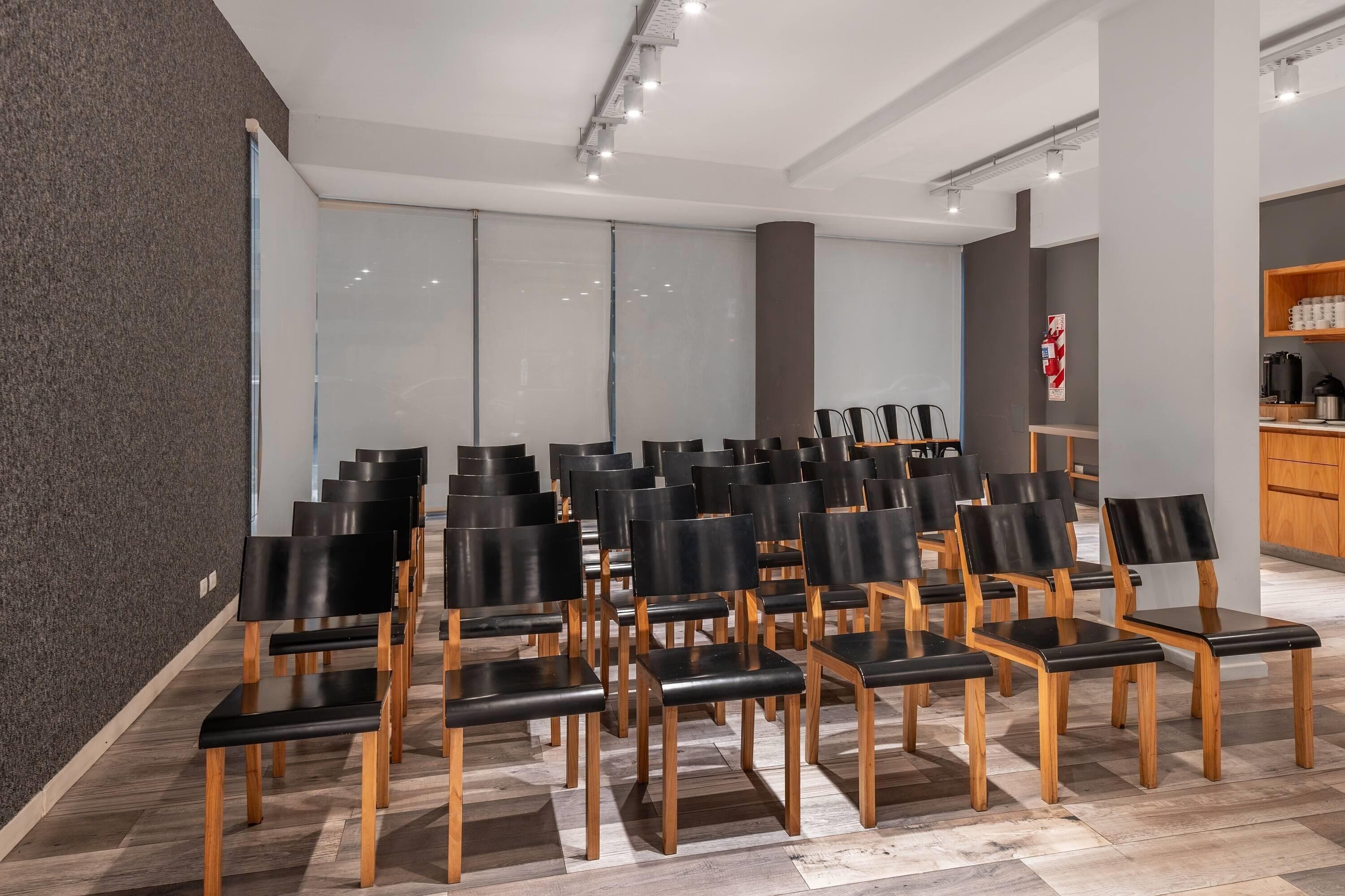 A conference room with rows of black chairs and wooden tables, large windows with blinds, and a kitchenette area.