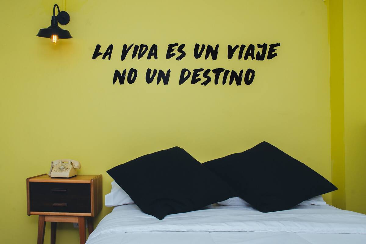 A room with a yellow wall, a bed with white bedding, black pillows, a wooden nightstand with a rotary phone, and a wall-mounted light fixture.