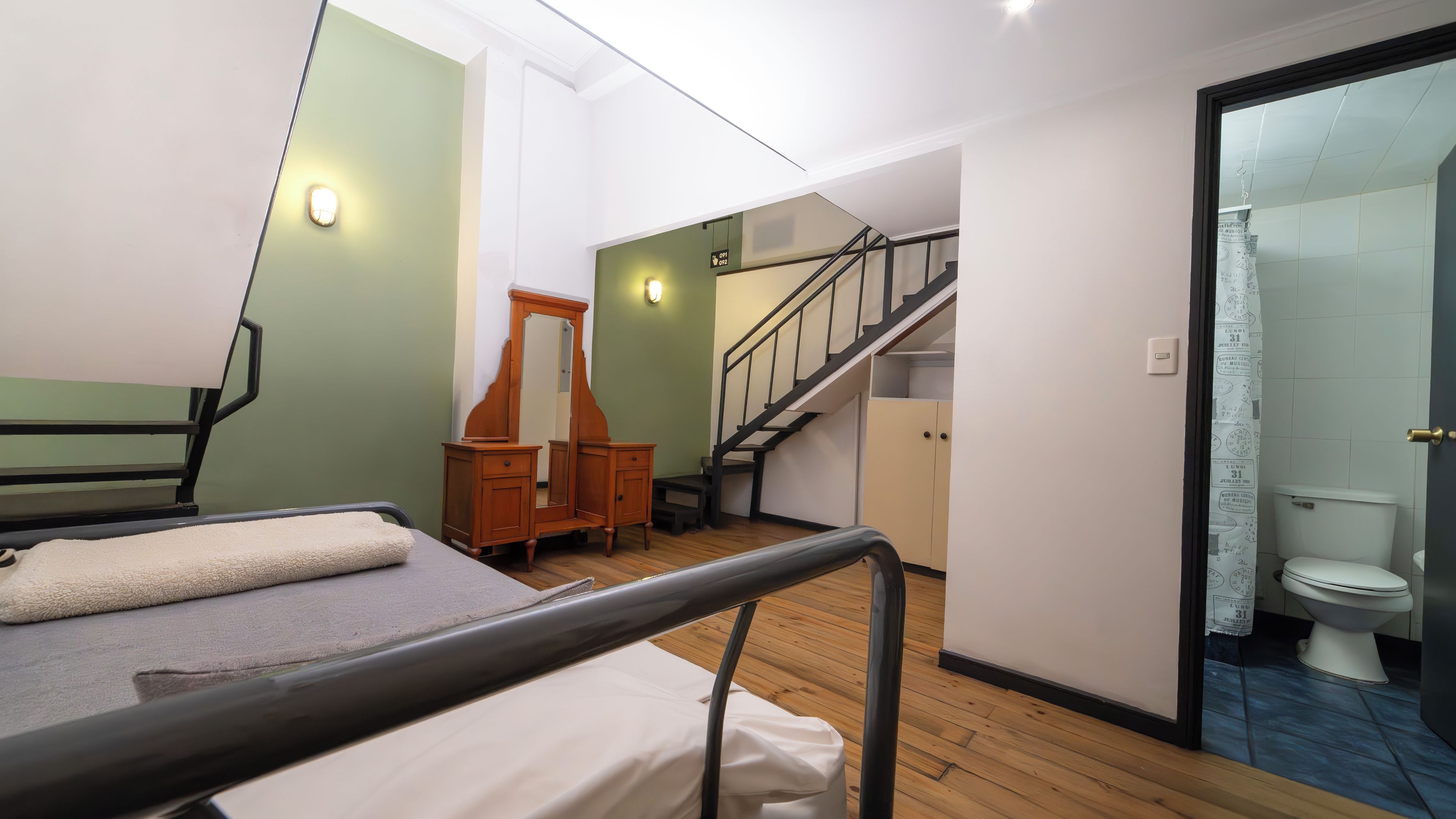 A room with a bed, a wooden dresser, a mirror, a staircase, and a bathroom with a toilet.