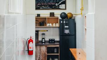 Cocina estrecha con paredes de azulejos blancos, una mesa de madera y un refrigerador negro. Un extintor de incendios está sujeto a la pared.