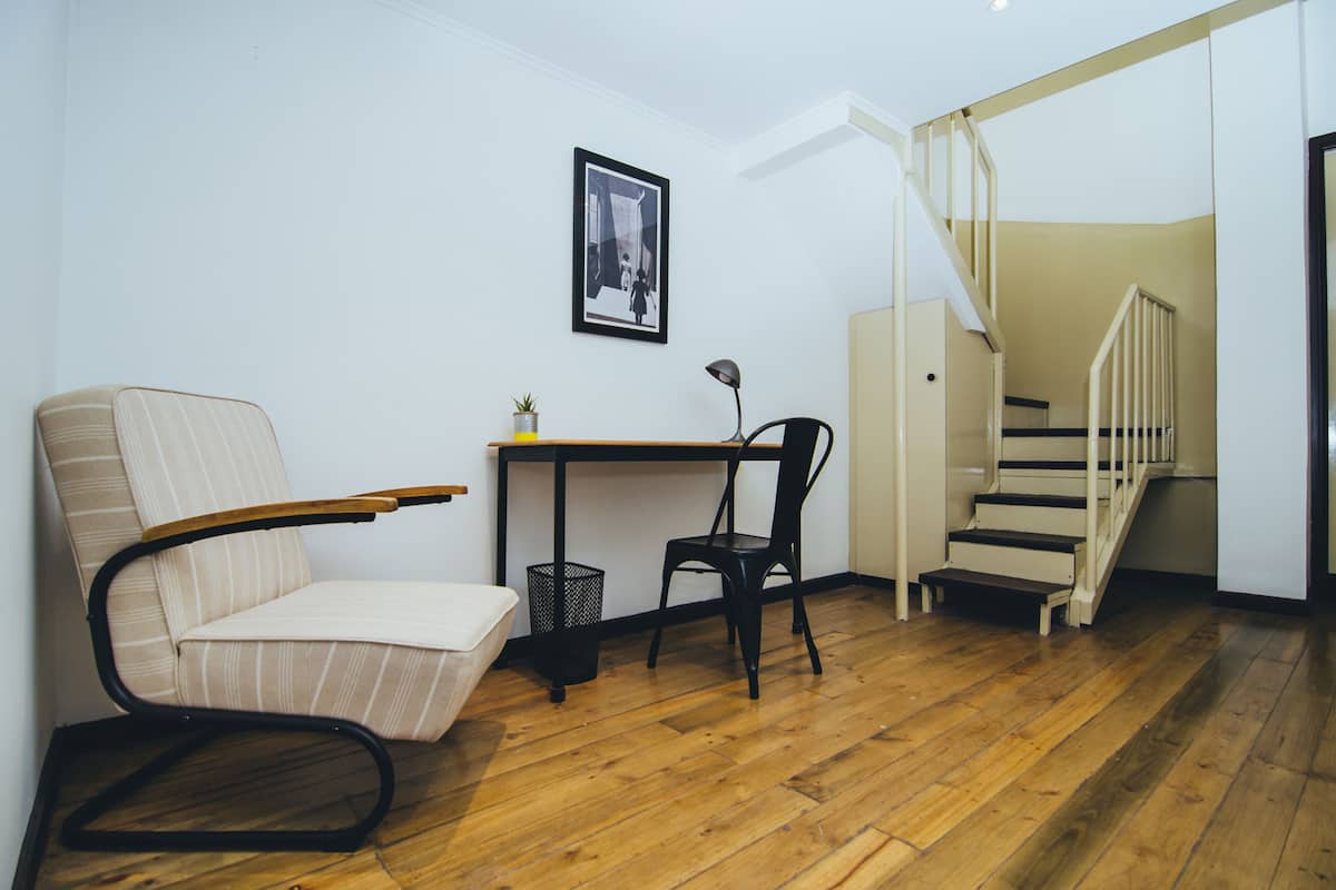 A room with a wooden floor, a striped armchair, a desk with a chair, a small potted plant, a framed picture on the wall, and a staircase leading to another level.