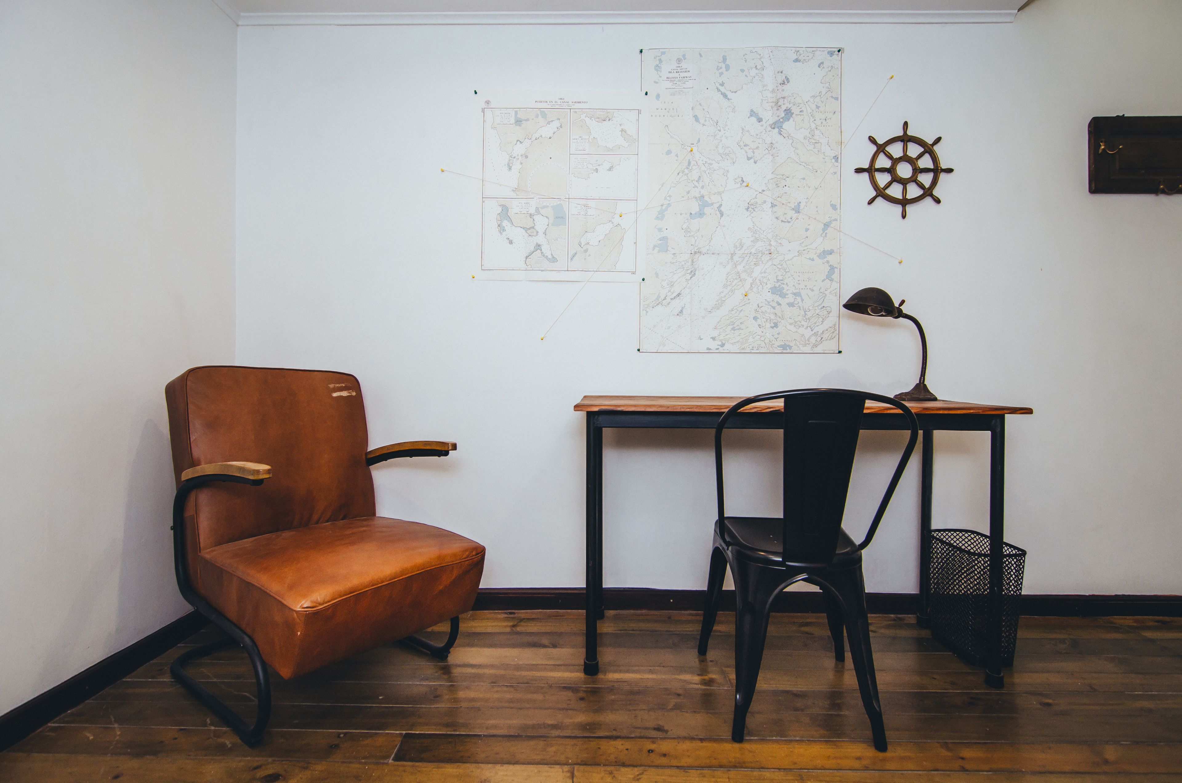 A room with a wooden floor, a brown leather chair, a black chair, a wooden desk, a ship's wheel decoration, and a map on the wall.