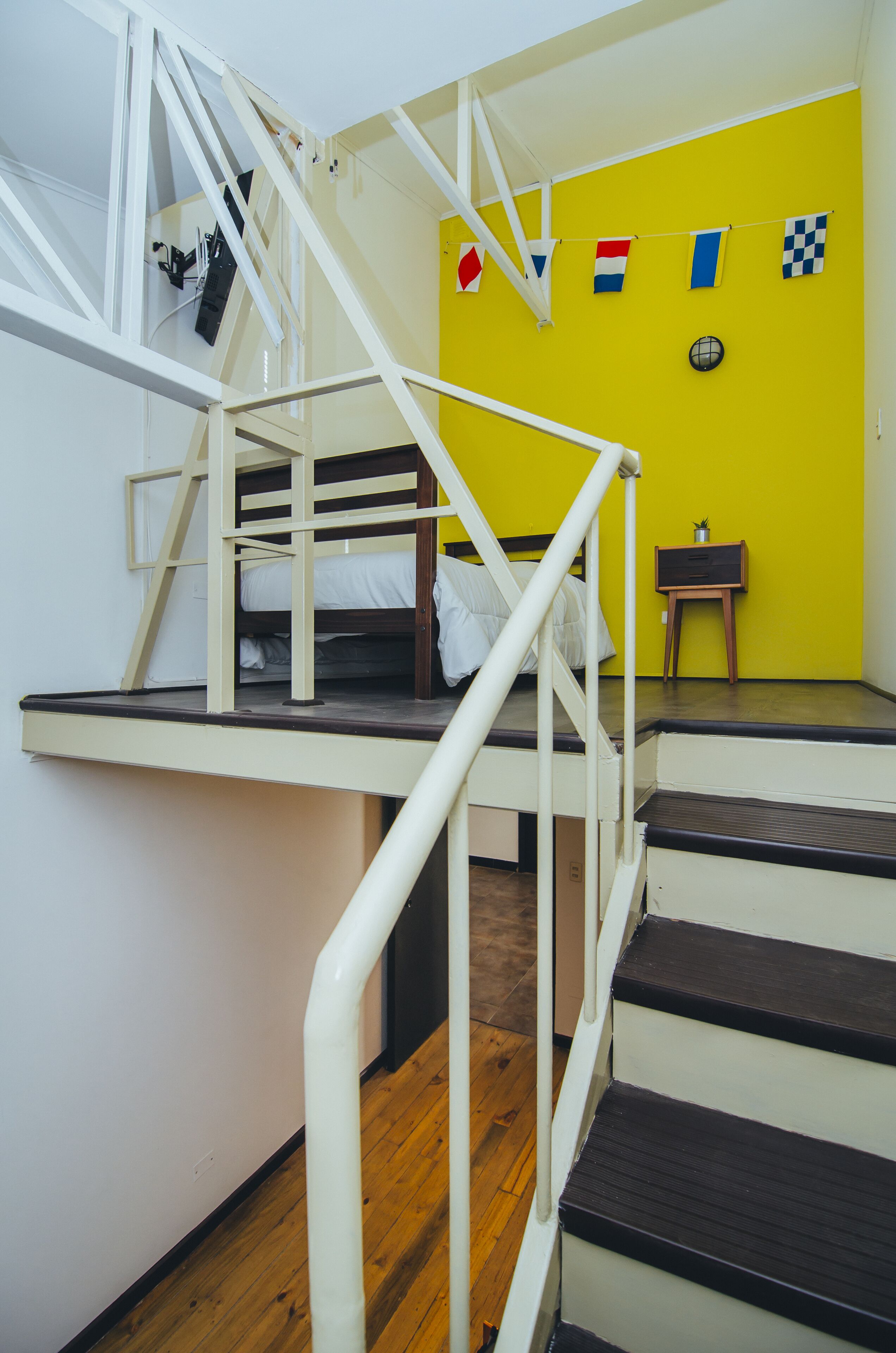 A bunk bed with a white railing, a small table with a plant, and a yellow wall with flags.