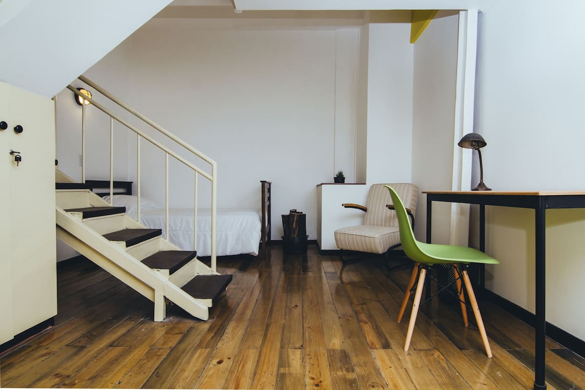 A room with a wooden floor, a bed with white bedding, a green chair, a small table with a lamp, and a staircase leading to another room.