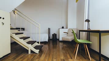 Une pièce avec un plancher en bois, un lit recouvert de draps blancs, une chaise verte, une petite table avec une lampe et un escalier menant à une autre pièce.