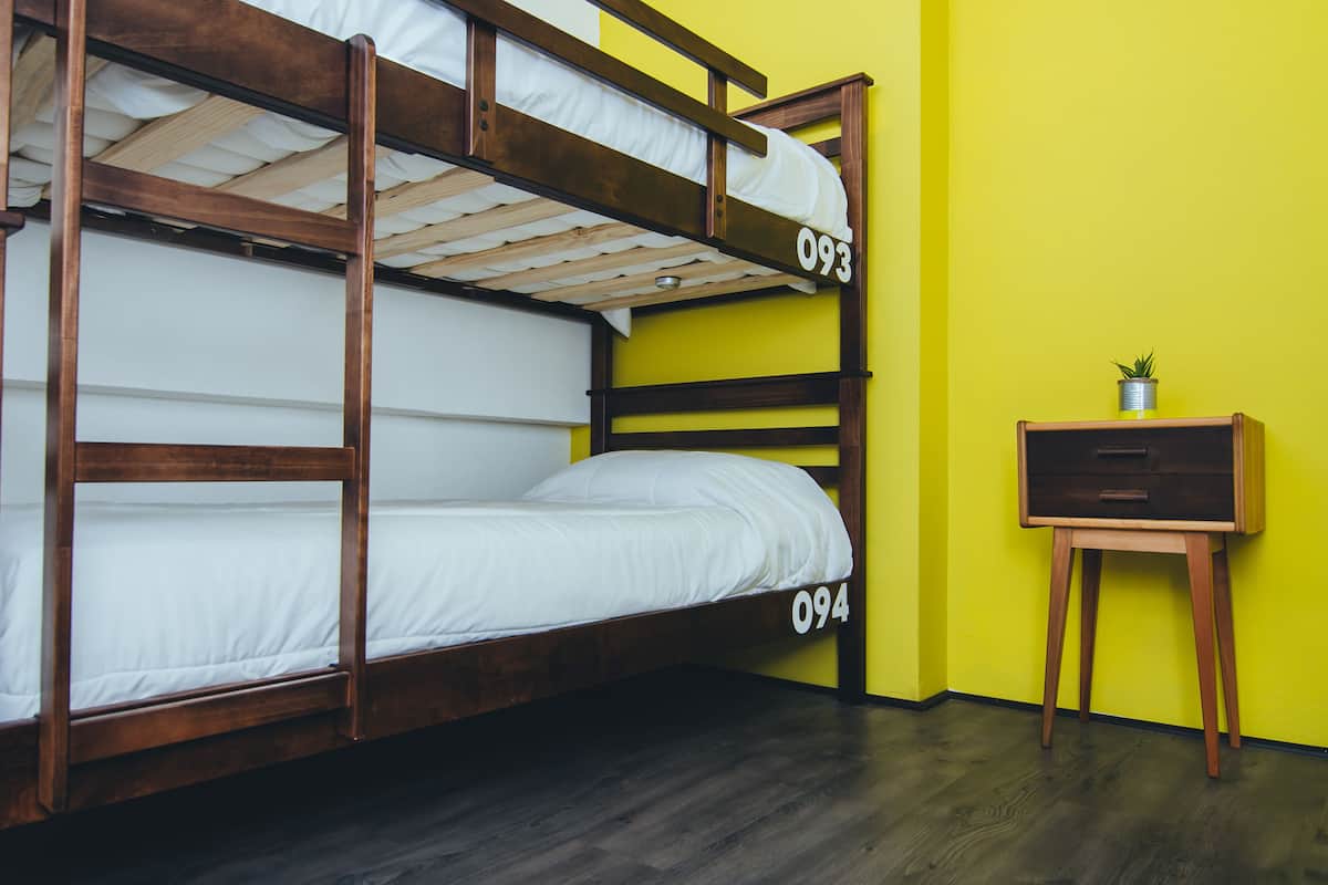 A bunk bed with numbers 093 and 094, a small wooden nightstand with a plant, and a yellow wall.
