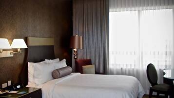 Habitación de hotel con dos camas, un escritorio con silla, una lámpara y vistas a la ciudad a través de amplias ventanas.