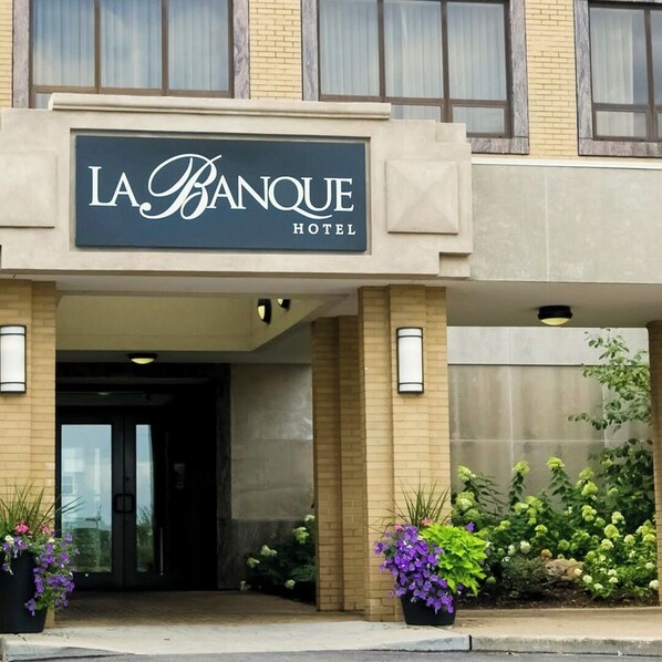 The entrance of La Banque Hotel with a sign above the door, potted plants, and a brick facade.