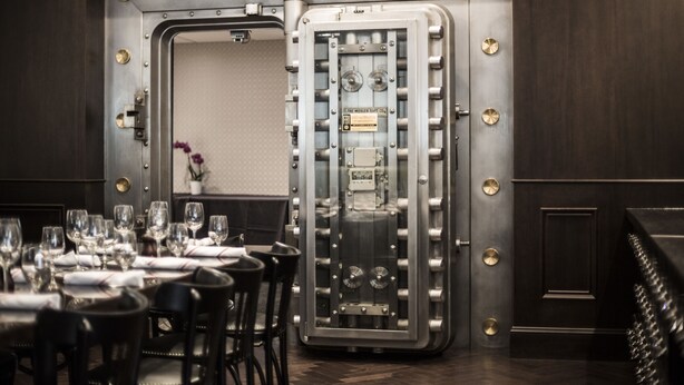 A room with a large safe deposit box, a dining table set with glasses and cutlery, and a vase with flowers. - La Banque Hotel (Homewood)