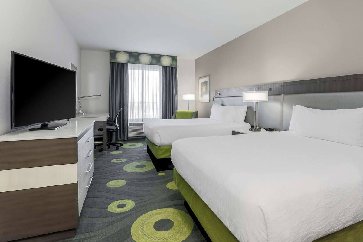 A hotel room with two beds, a desk with a TV, a chair, a lamp, and a window with curtains.