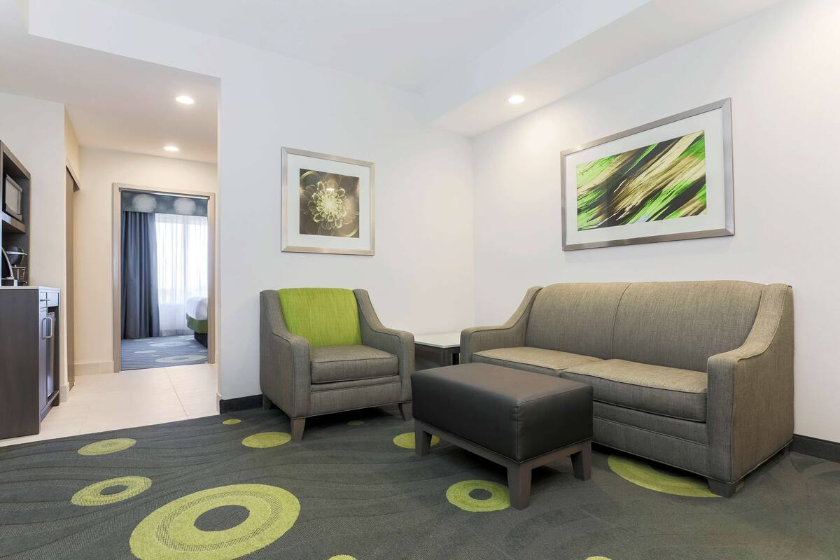 A modern hotel room with a grey sofa, a green armchair, and a black ottoman. There are two framed artworks on the walls and a kitchen area in the background.