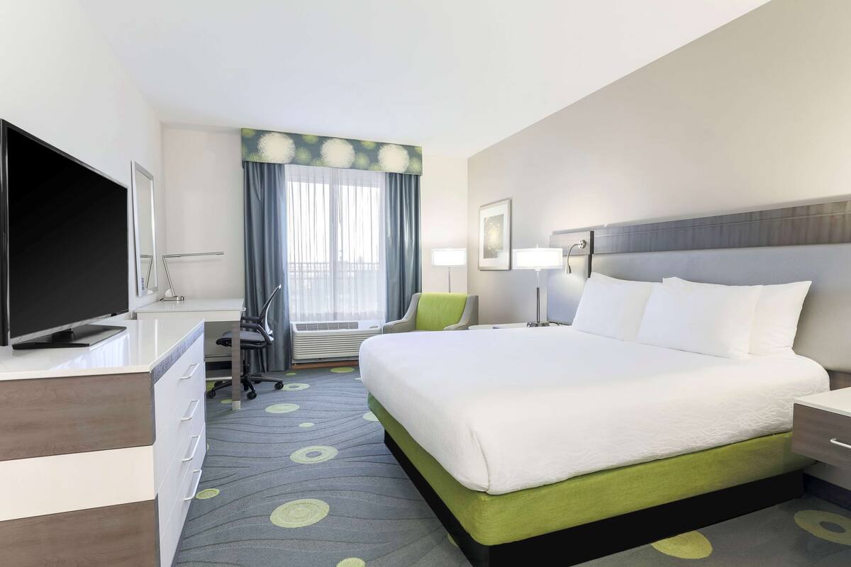A hotel room with a large bed, a desk with a flat-screen TV, a chair, a desk lamp, and a window with curtains.