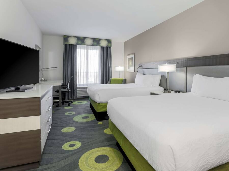 A hotel room with two beds, a desk with a flat-screen TV, a chair, a lamp, and a window with curtains.