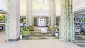 Un hall d’entrée spacieux, avec un canapé vert, une table basse et un tapis à motifs. On y trouve des rideaux ornés de motifs de feuilles, un comptoir d’accueil et une grande fenêtre donnant sur un paysage verdoyant.