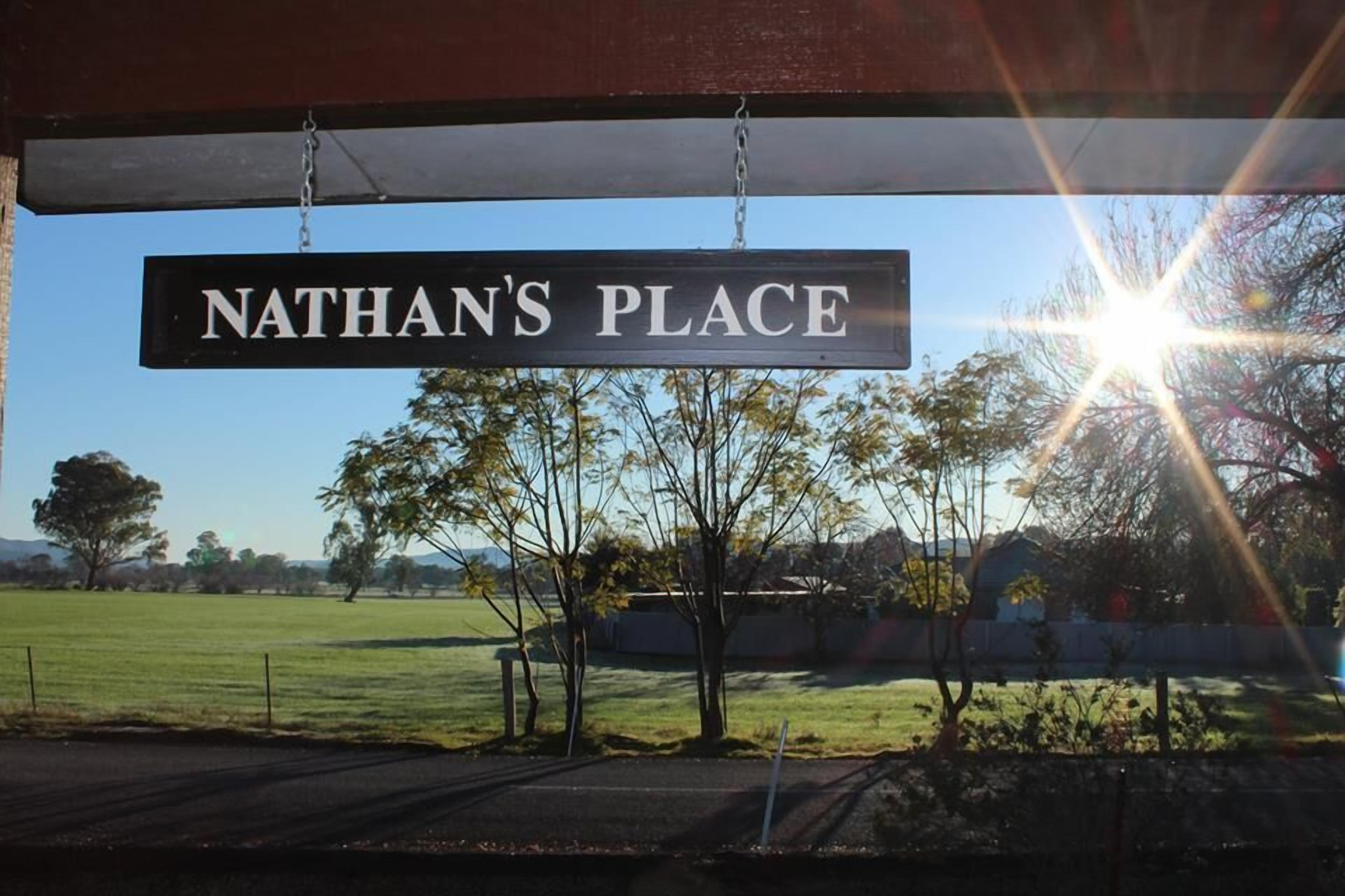 Un letrero con la leyenda 'NATHAN'S PLACE' colgado debajo de una estructura, con vista despejada a un campo cubierto de césped y árboles al fondo.