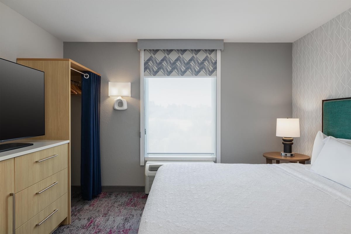 A hotel room with a bed, a TV, a wardrobe, a window with a view, and a bedside table with a lamp.
