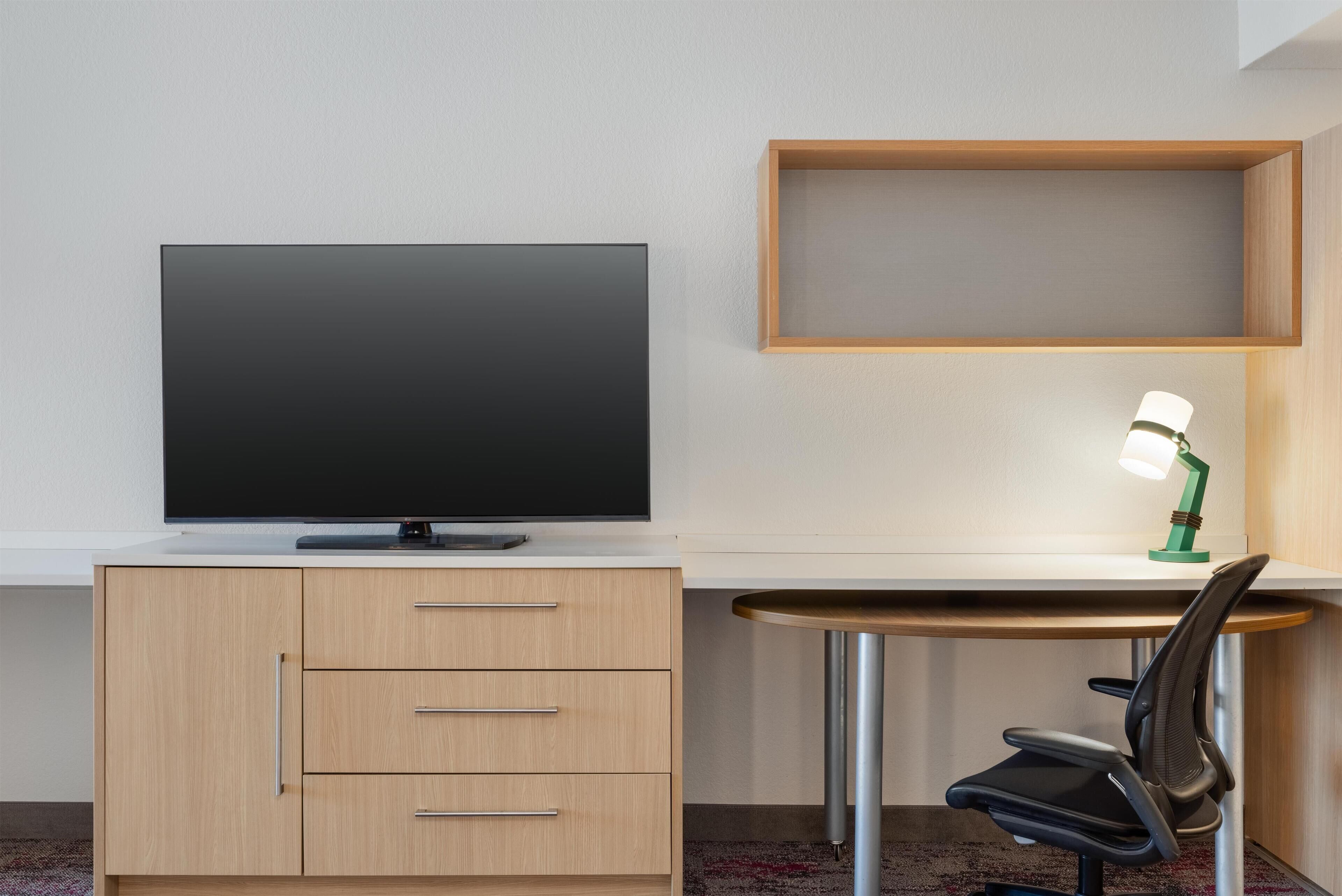 Une chambre moderne avec un téléviseur à écran plat posé sur un meuble en bois, un bureau avec une chaise et une lampe de bureau verte.
