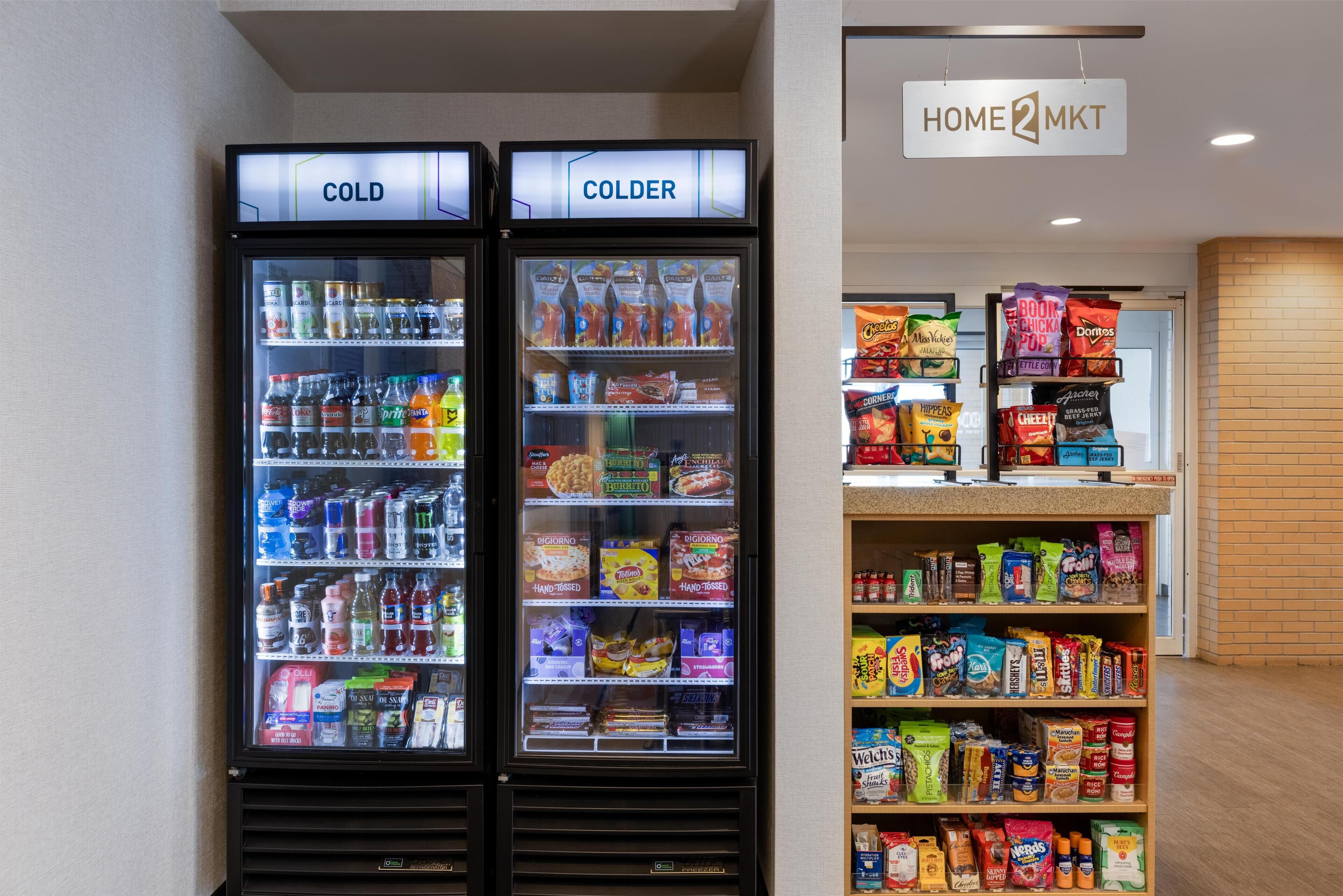 Two refrigerated beverage dispensers labeled 'COLD' and 'COLDER' with a variety of drinks and snacks. A sign above reads 'HOME 2 MKT'.