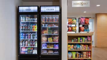 Two refrigerated beverage dispensers labeled 'COLD' and 'COLDER' with a variety of drinks and snacks. A sign above reads 'HOME 2 MKT'.