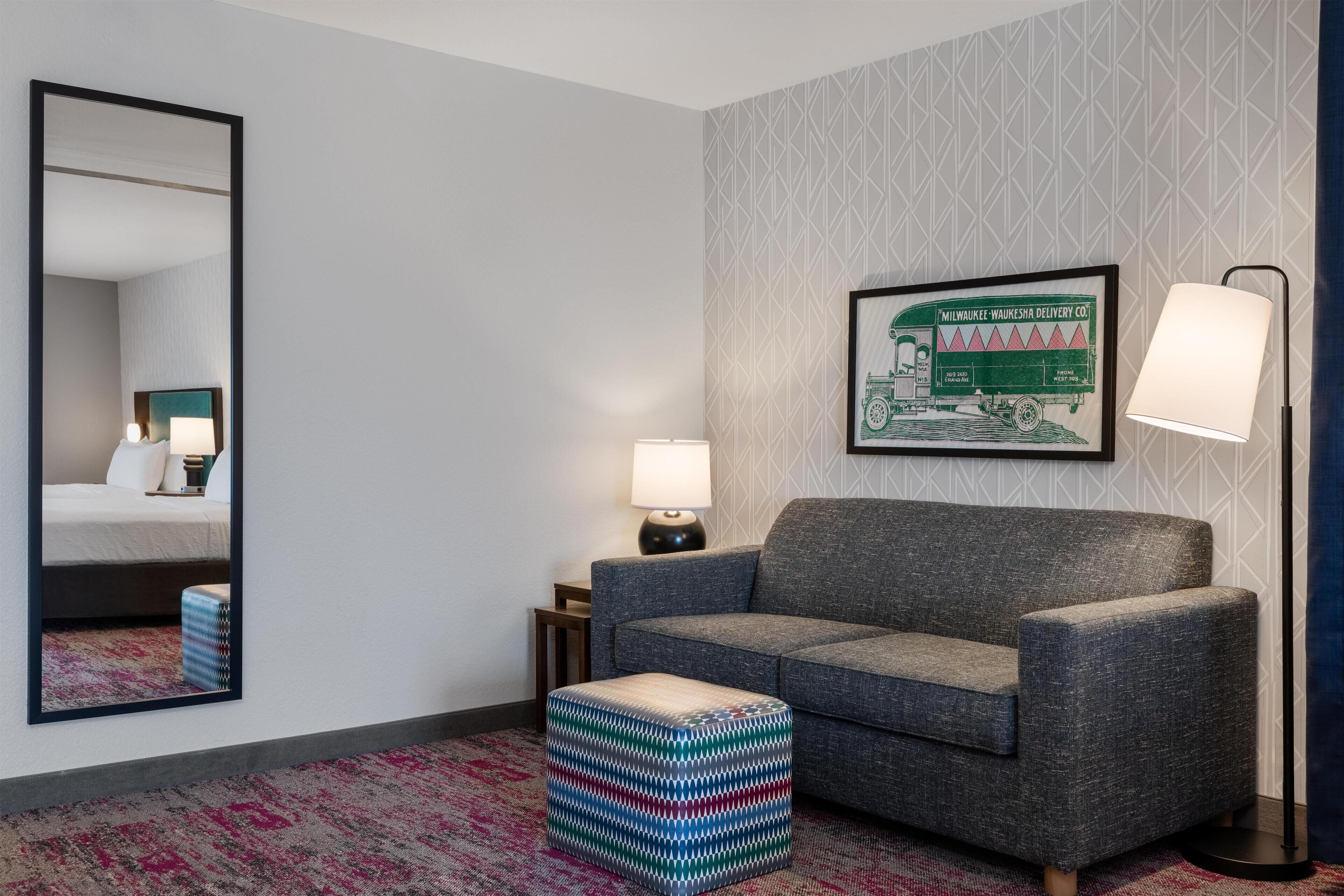 A hotel room with a grey sofa, a patterned ottoman, a framed picture on the wall, a mirror, and a lamp.