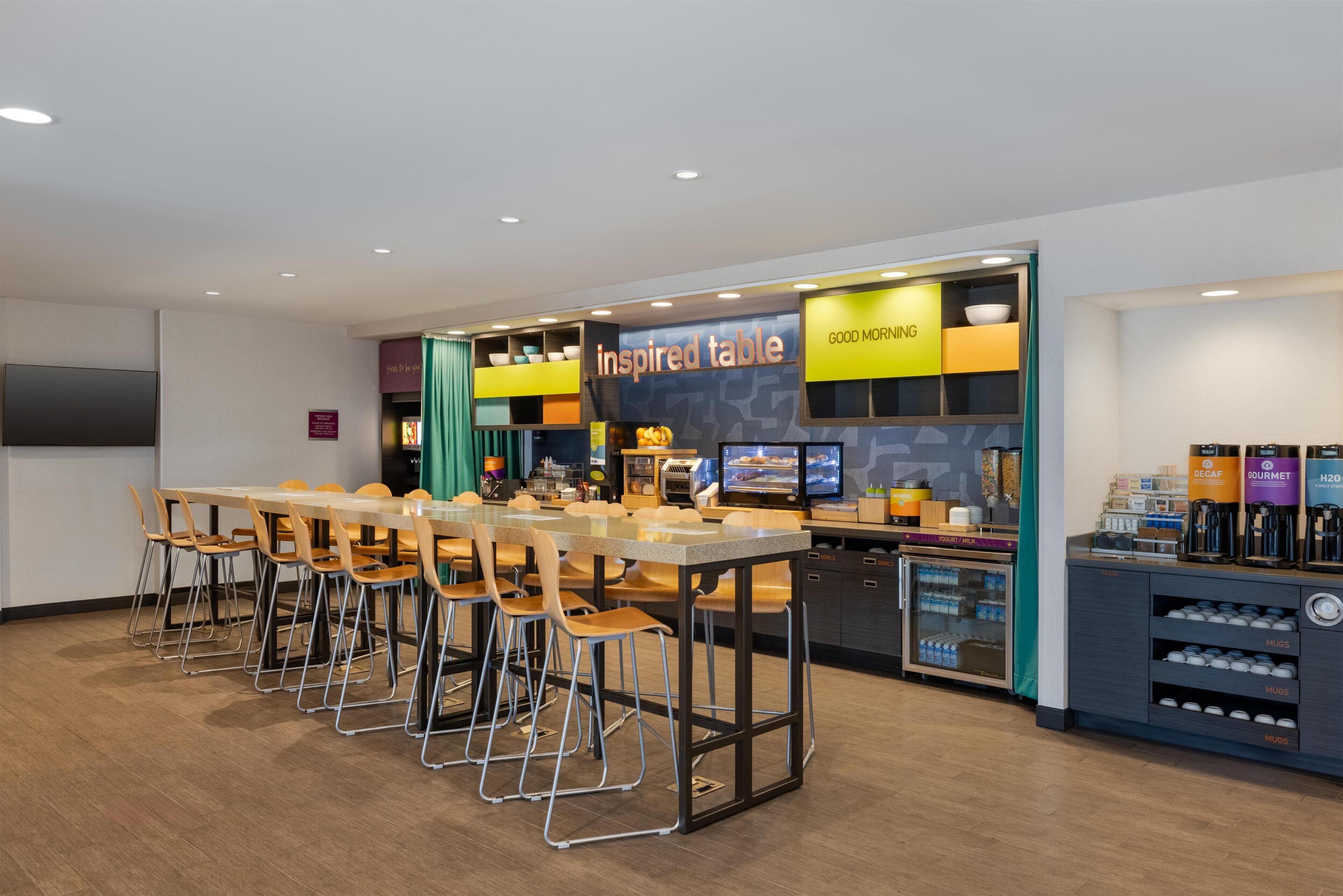 A modern cafe with a long communal table and bar stools, a coffee station, and a menu board.