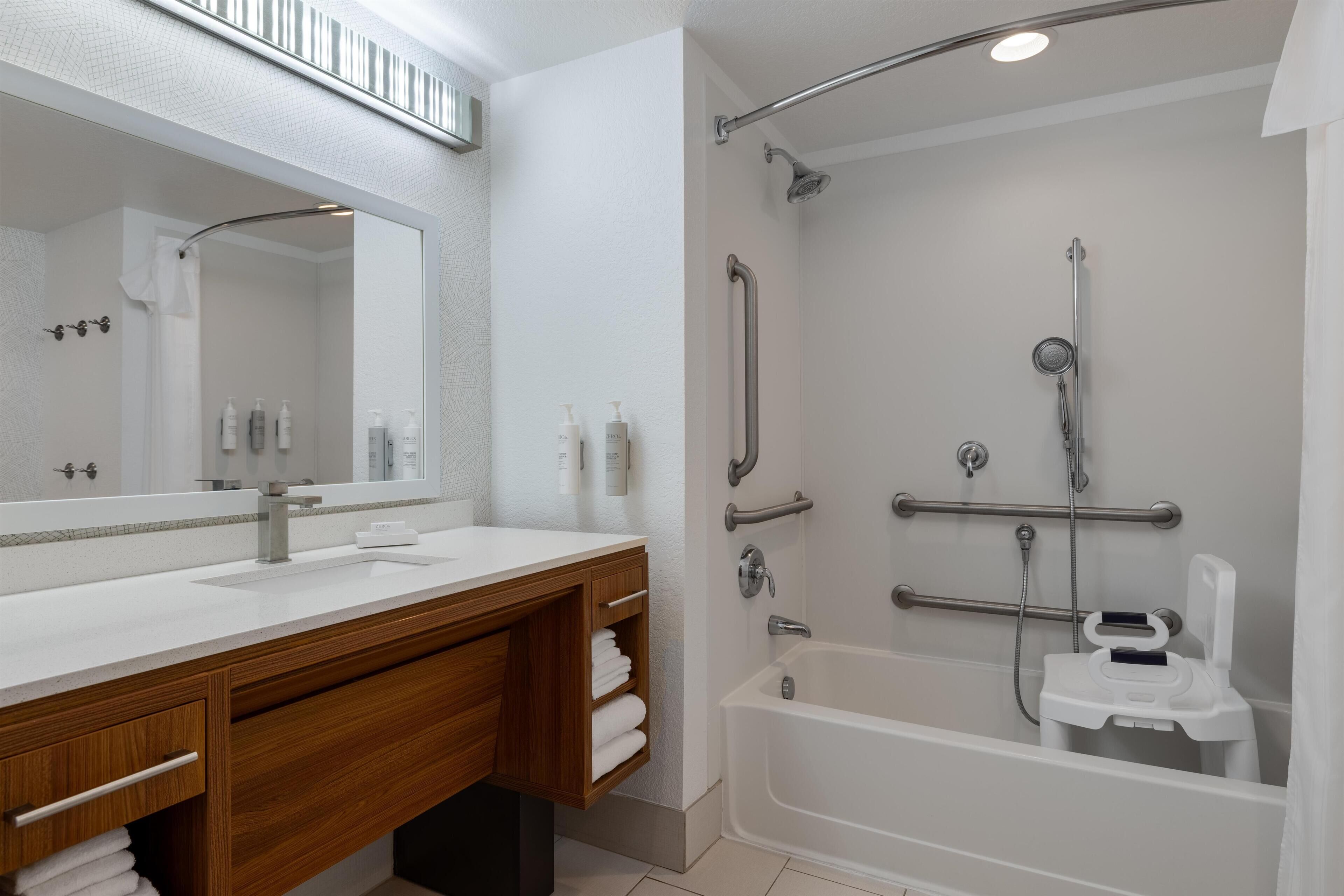 Un baño moderno con un espejo grande, lavamanos blanco, tocador de madera y ducha a ras de suelo con barra de apoyo.