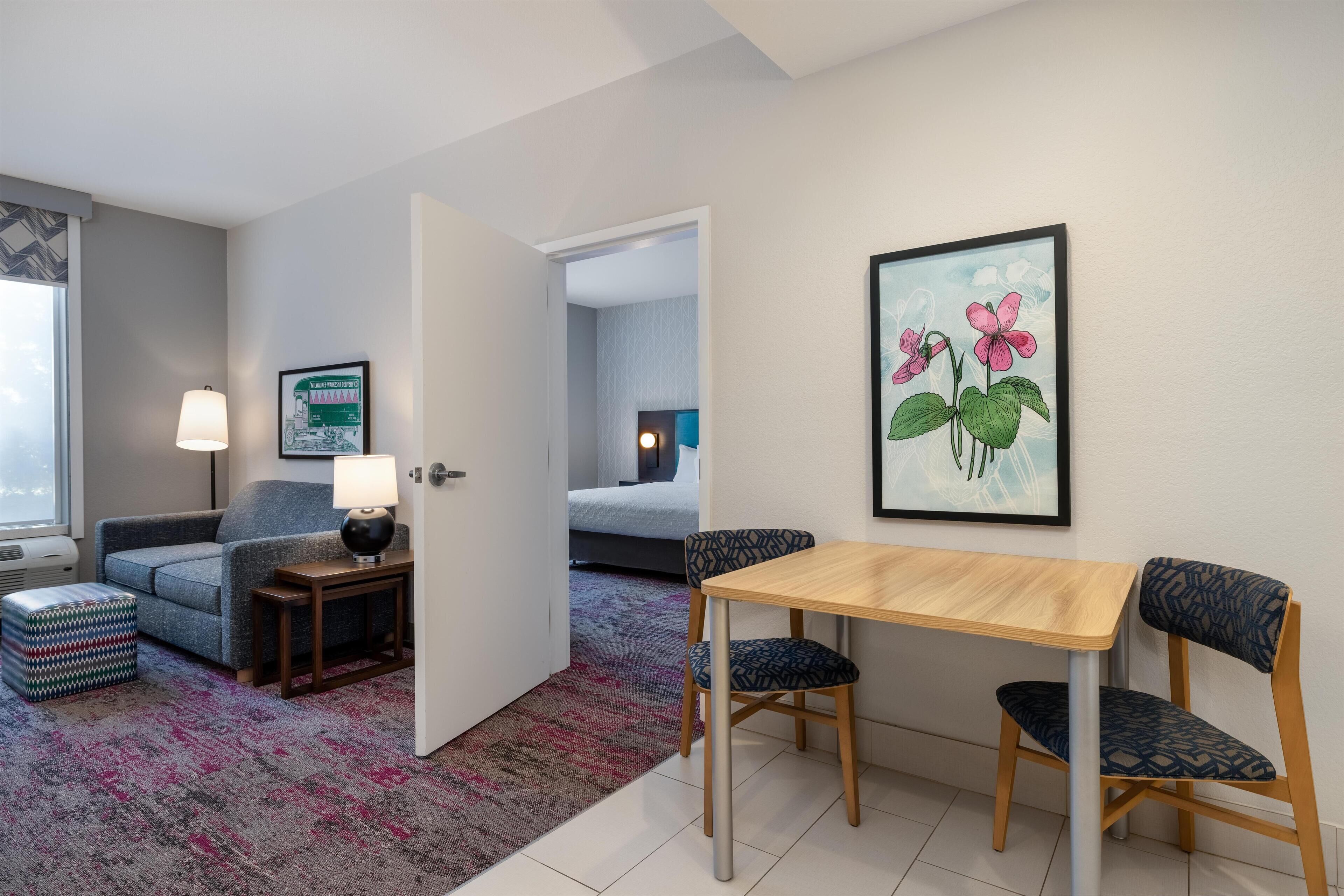 A modern hotel room with a sofa, a small table with a lamp, a dining table with chairs, a colorful rug, and a framed picture on the wall.