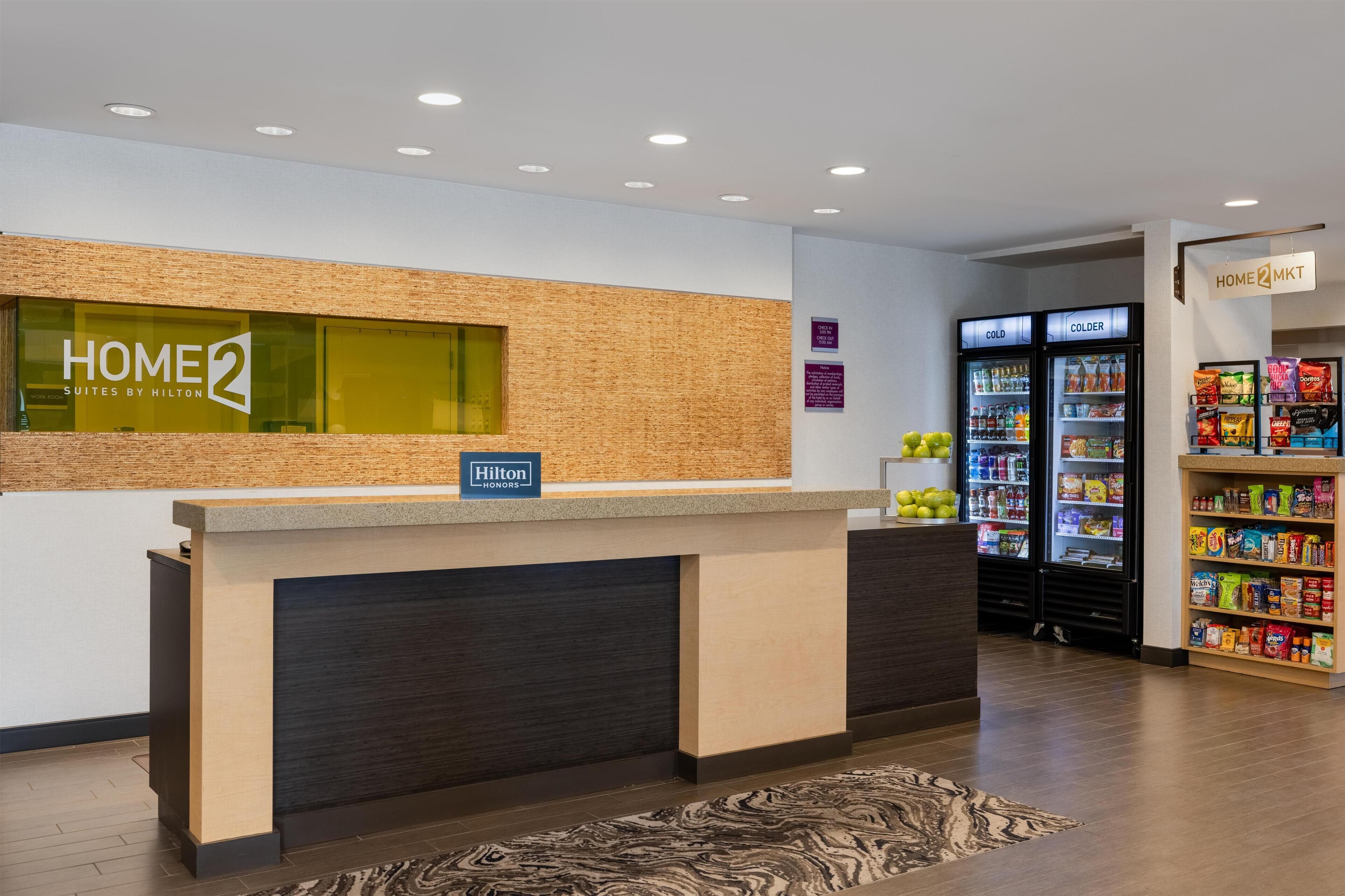 En hotellobby med en receptionsdisk, en skylt med texten ”HOME 2 SUITES BY HILTON” och en försäljningsautomat märkt ”HOME 2 MKT”.