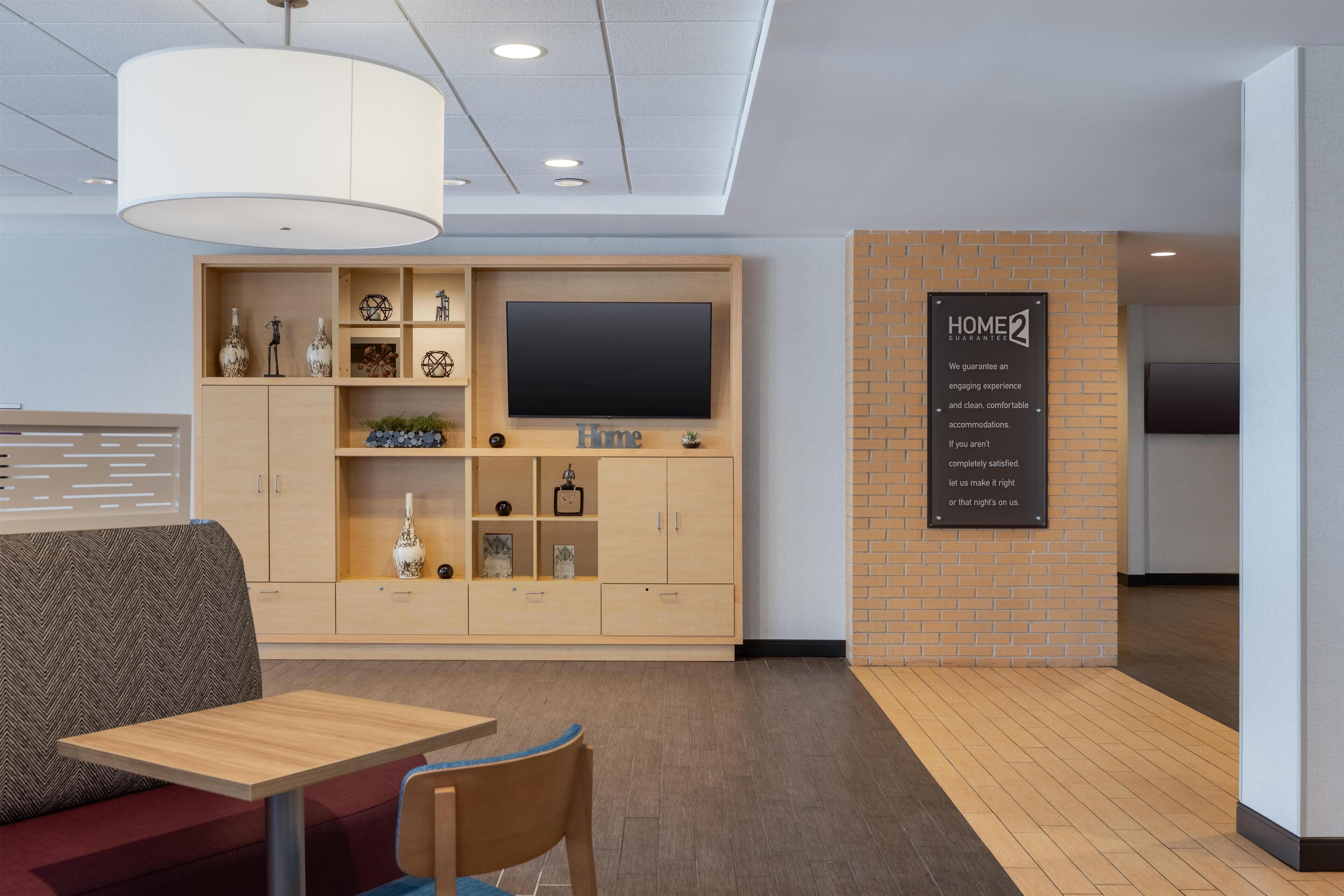 A modern hotel room with a wooden cabinet, a flat-screen TV, a 'Home Guarantee' sign, a sofa, and a table.
