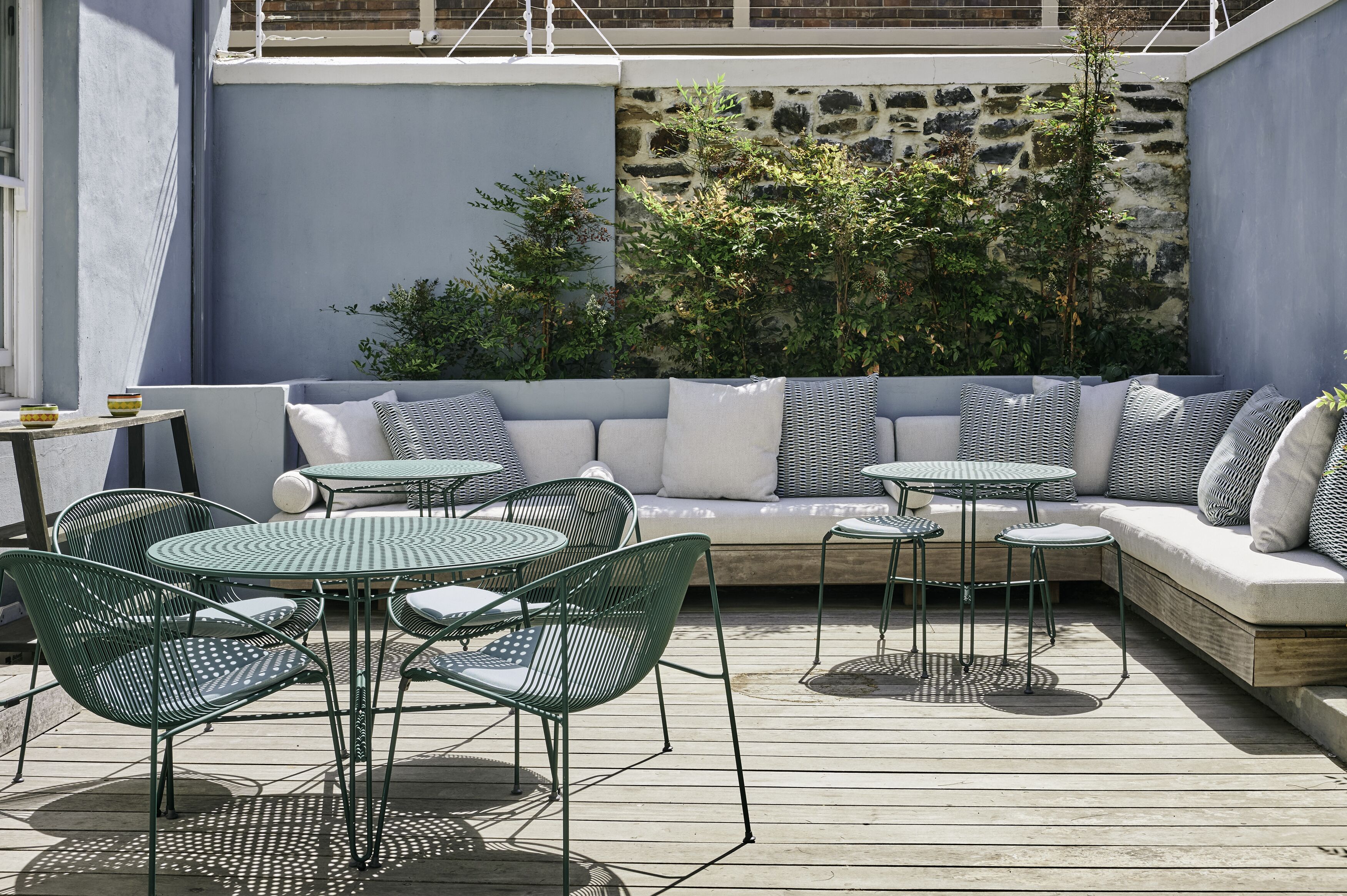 An outdoor seating area with a sofa, round tables, and chairs, surrounded by greenery and a stone wall.