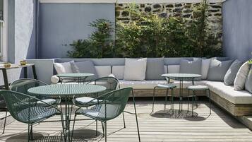 An outdoor seating area with a sofa, round tables, and chairs, surrounded by greenery and a stone wall.