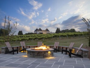 A stone fire pit with chairs, a vineyard in the background, and a large building with a distinctive roof. - Hampton Inn & Suites Braselton (Braselton)
