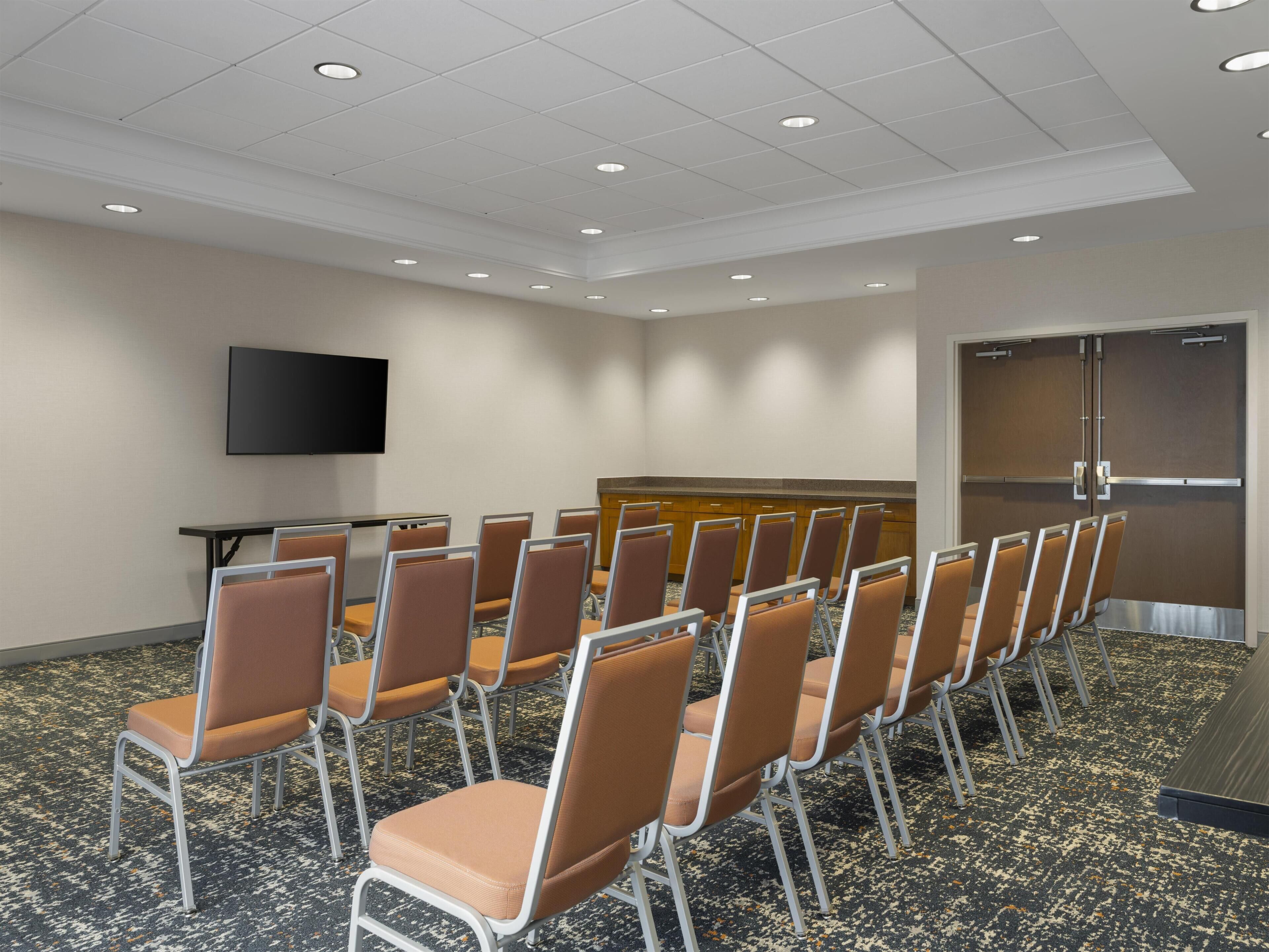 Une salle de conférence avec des rangées de chaises, un téléviseur à écran plat, un pupitre et une porte.